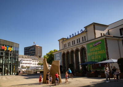 Apollo Theater. An einem sonnigen Tag entstanden. Links davon befindet sich der Laden MyToys und davor befinden sich Menschen, die sich am Springbrunnen die Füße abkühlen.